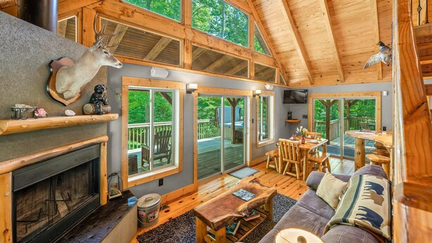 Una cabaña espaciosa con techo alto, vigas de madera, chimenea, zona de comedor y ventanas grandes que ofrecen vistas al exterior.
