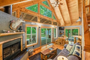 Una cabaña espaciosa con techo alto, vigas de madera, chimenea, zona de comedor y ventanas grandes que ofrecen vistas al exterior.