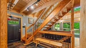 Una escalera de madera con un banco, una puerta oscura y vistas al exterior, con vegetación.