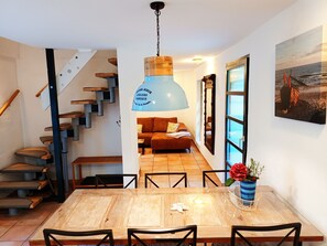 A dining area with a wooden table and chairs, a blue pendant light, a staircase, a sofa, and a large window with a view. - Apartment Hühnergott - Kölp - forest and sea(h)r (Loddin)