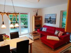 A living room with a red sofa, a wooden coffee table, a bookshelf, and a dining area with a table and chairs. - Apartment Hühnergott - Kölp - forest and sea(h)r (Loddin)