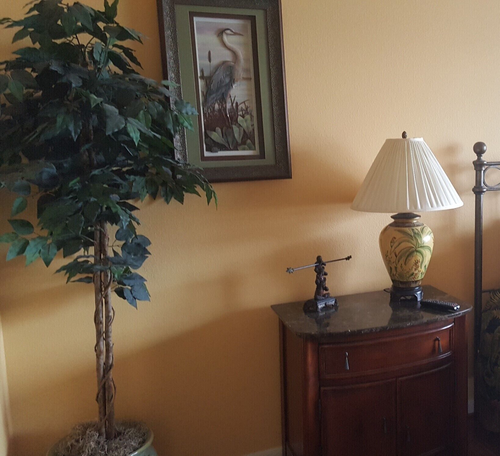 A room with a potted plant, a framed picture on the wall, a lamp on a wooden side table, and a remote control on the table.