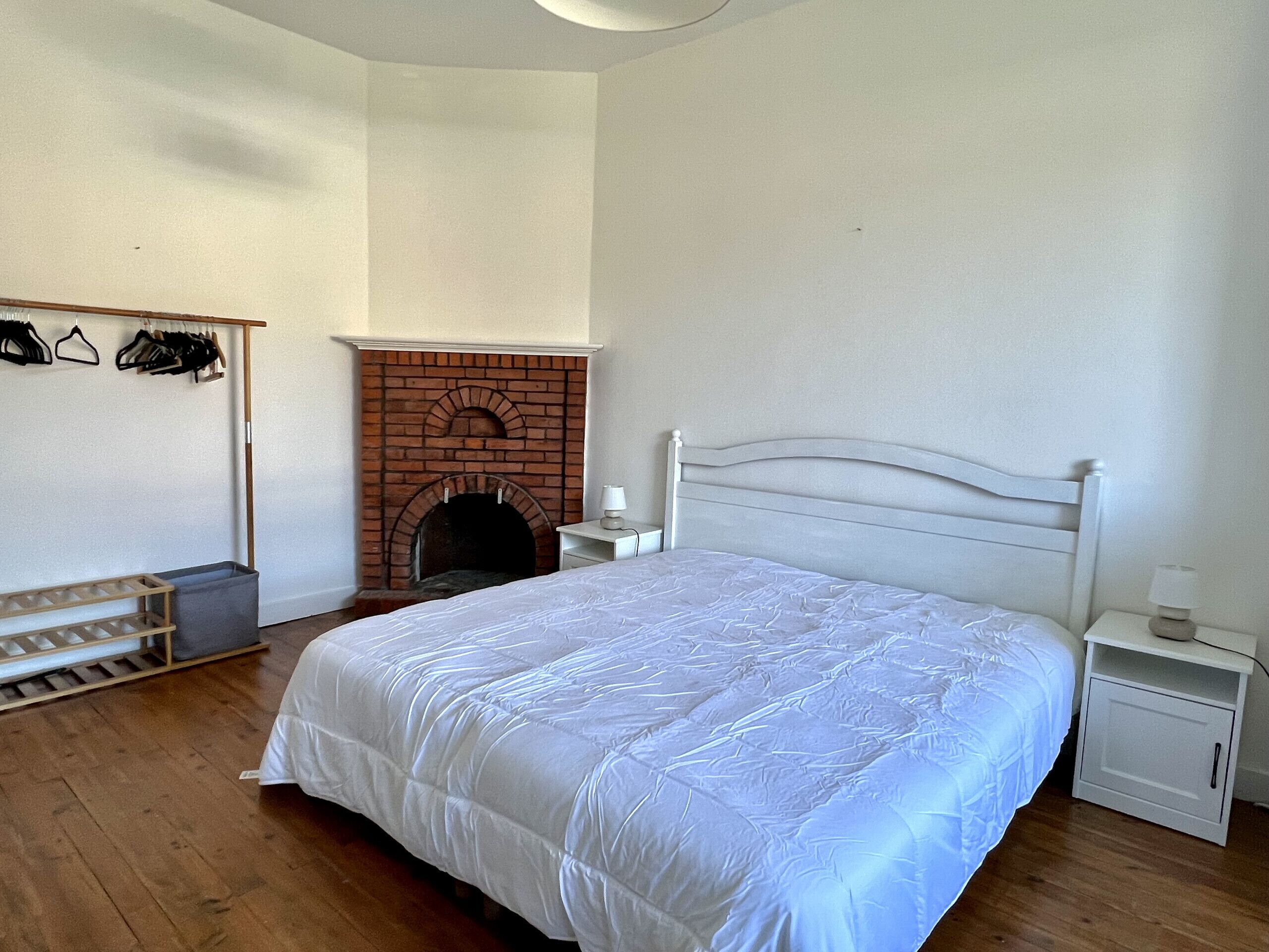 Un dormitorio con una cama blanca, una chimenea de ladrillo, un banco de madera y una mesita de noche blanca con una lámpara.