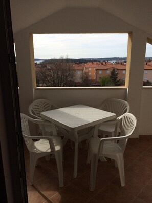 Un balcón con una mesa y sillas blancas, vistas a edificios y suelo de baldosas.