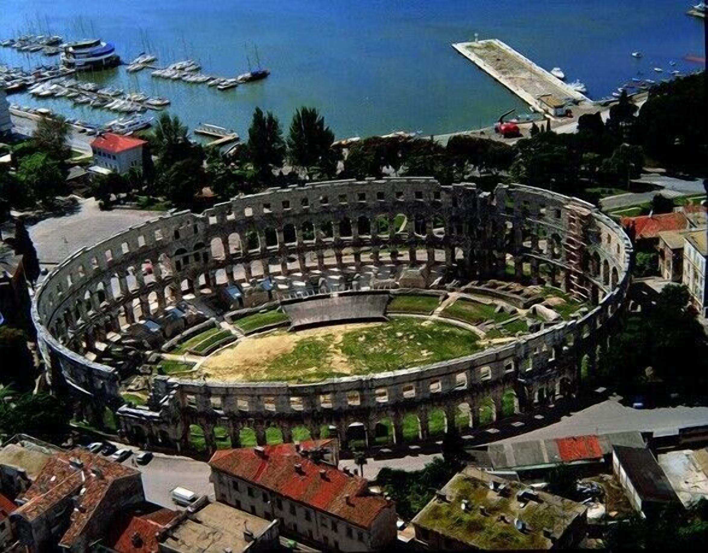 Ein uraltes, kreisförmiges Amphitheater, umgeben von einer Marina mit Booten und Blick auf die Küste.