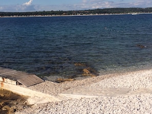 Una playa de guijarros con un muelle de madera que se adentra en aguas cristalinas y azules.