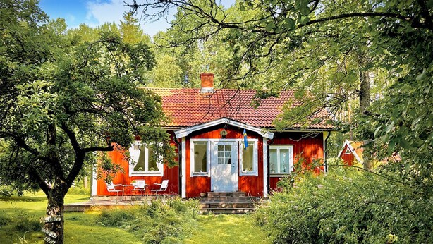 Une maison en bois rouge, avec un toit recouvert de tuiles, des fenêtres à cadre blanc et un porche à l’avant, orné de chaises blanches.