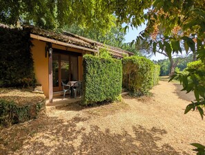 Un bâtiment rustique avec un toit de chaume, un patio couvert avec des chaises et un chemin de gravier menant à l’entrée.