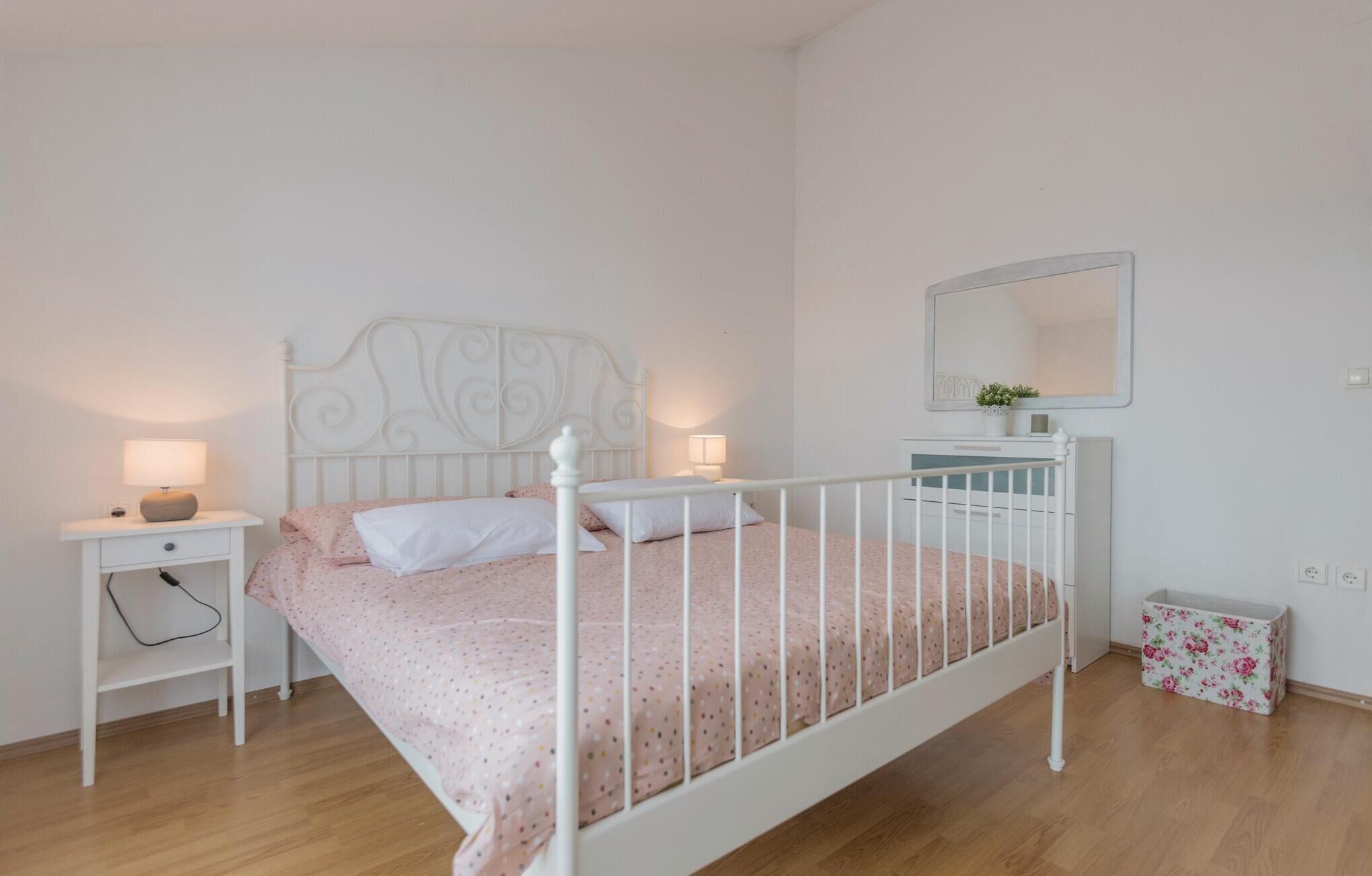 A bedroom with a white metal bed frame, a bedside table with a lamp, a dresser, and a mirror on the wall.