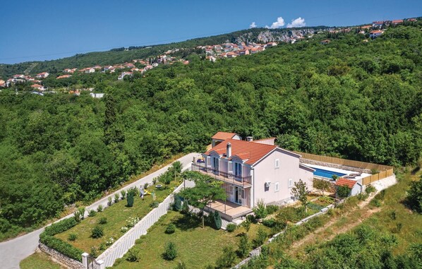 A modern two-story house with a swimming pool, surrounded by lush greenery and a hillside village in the background.