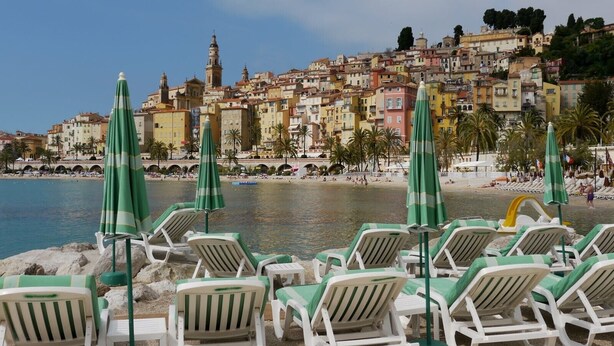 A beach with rows of white lounge chairs and green umbrellas, a clear view of a colorful coastal town in the background. - Beautiful for 4 people, town centre, 100 m from the beaches, near the shops (Menton)