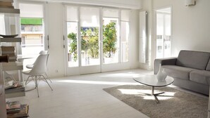 A modern living room with a grey sofa, a white chair, a glass coffee table, and a large sliding glass door leading to a balcony with greenery. - Beautiful for 4 people, town centre, 100 m from the beaches, near the shops (Menton)