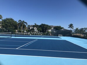 Ein Tennisplatz im Freien mit blauem Belag, umgeben von Bäumen und einem Zaun.