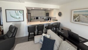 A modern kitchen with white cabinets, a central island with bar stools, and a dining area with a sofa and a laptop on a table.