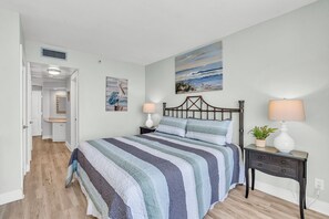 A bedroom with a bed, bedside lamps, a nightstand, a painting, and a door leading to another room.