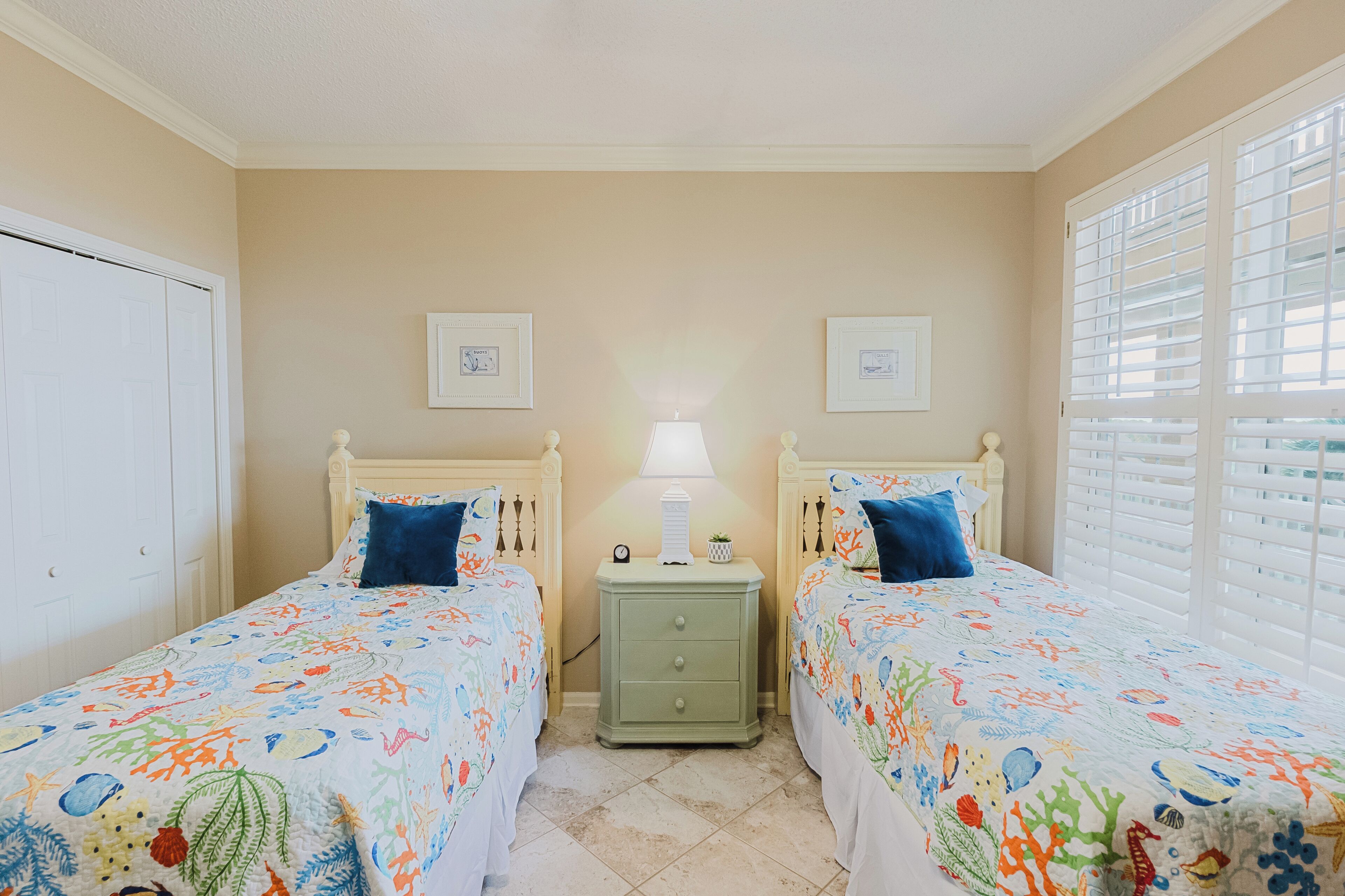 A bedroom with two beds, a nightstand with a lamp, and a window with blinds.