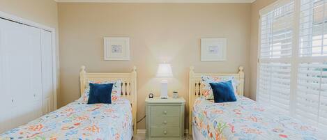 A bedroom with two beds, a nightstand with a lamp, and a window with blinds.