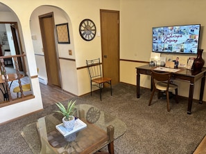 A furnished room with a wooden desk, a chair, a potted plant, a clock, and a television displaying a website.