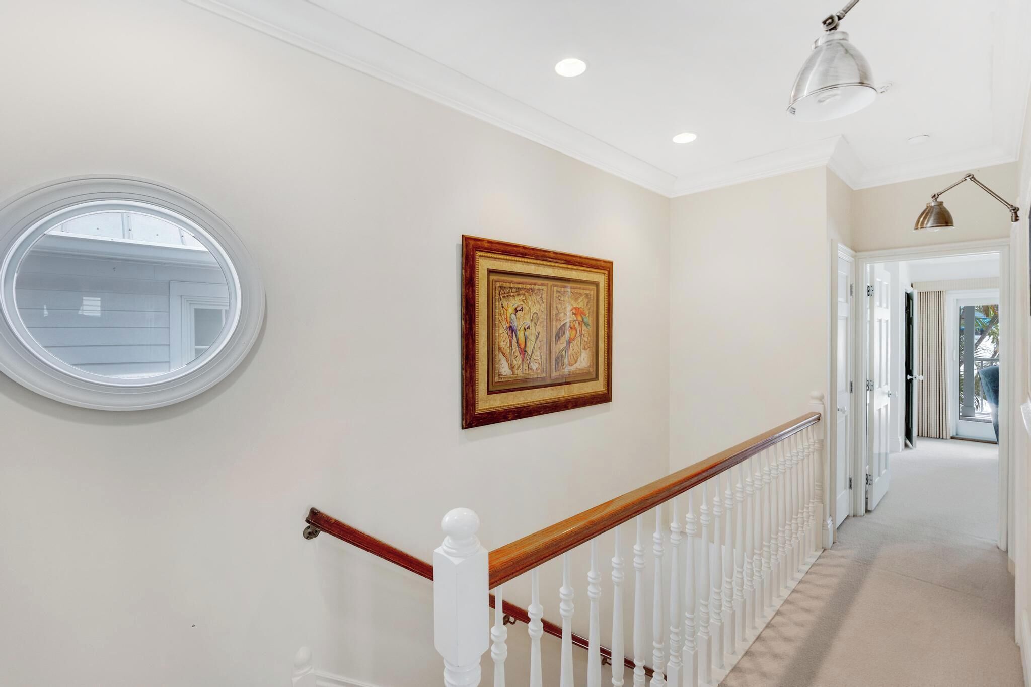 A hallway with a white railing, a round window, and a framed artwork on the wall.