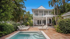 Un espace piscine avec des chaises longues, un bâtiment à deux étages doté d’un balcon et une végétation luxuriante.