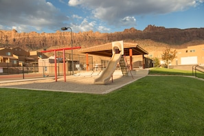 A playground with a slide and swings, surrounded by a fenced area, with buildings and a mountain in the background. - Desert Solitude Rim Vista 5A3 (Moab)
