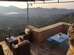 A terrace with wicker furniture, a table, and chairs, overlooking a mountainous landscape.