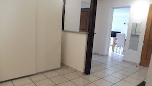 Un couloir avec un sol carrelé, un cadre de porte en bois, et une cuisine visible au fond.