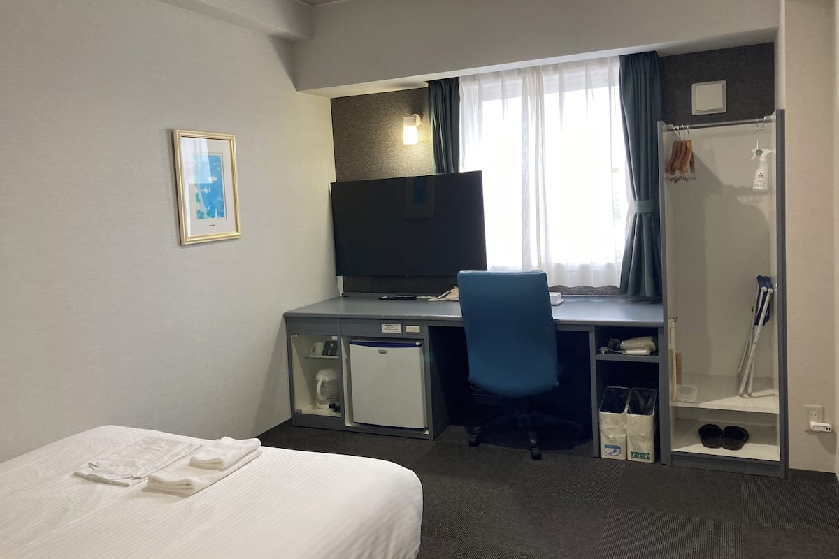 A hotel room with a bed, a desk with a computer, a television, a shower, and a window with curtains.