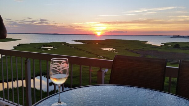 A glass of white wine on a table with a view of a sunset over a body of water and green landscape. - Bay Front Condo-2br/2ba , Pool, Hot Tub, Lr Balcony W/ Beautiful Sunsets, WIFI.  (Ocean City)