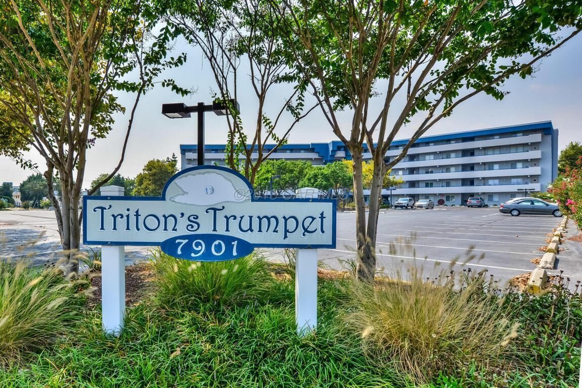 Skilt for Triton's Trumpet med adressen 7901, omgivet af grønt og en parkeringsplads med en bygning i baggrunden.