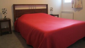 Una camera da letto con un letto in legno, una coperta rossa, un comodino con una lampada e una parete con un quadro floreale.