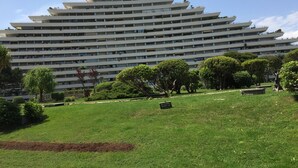 Un immeuble à plusieurs étages doté d’une architecture moderne, entouré d’un espace verdoyant bien entretenu, avec des arbres et un banc en pierre.