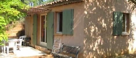 A small, rustic building with a tiled roof and green shutters, a bench, and a table outside.