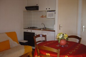 A compact kitchen with a stove, sink, and microwave. There's a dining table with a red tablecloth and a vase of flowers. A sofa with an orange cushion is in the foreground.