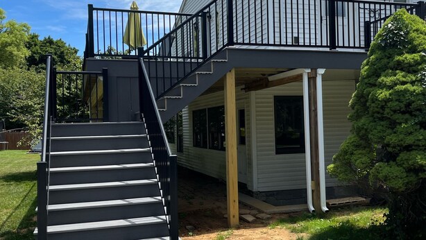 Een tweelaags huis met een zwarte trap die leidt naar een balkon, een klein terras en een hond in de voortuin.