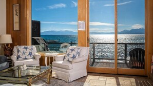 Une pièce chaleureuse avec vue sur la mer, comprenant un fauteuil agrémenté de coussins à motifs d’étoiles, une table basse en verre et une porte en bois donnant sur une terrasse extérieure.