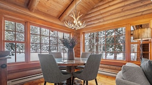 Ein Interieur einer Holzhütte mit Esstisch, Stühlen, einer Kronleuchter und großen Fenstern mit Blick auf schneebedeckte Bäume.