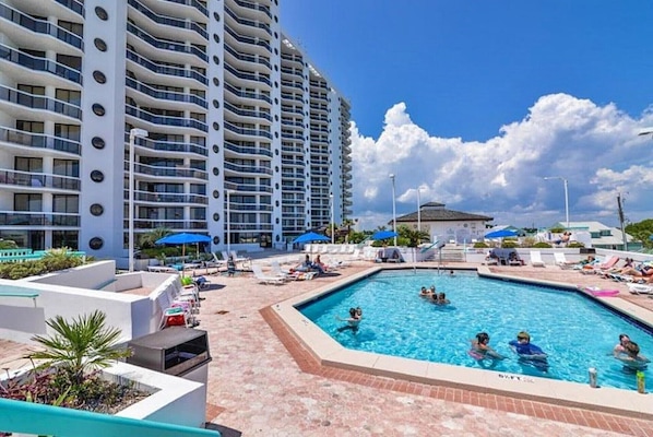 A swimming pool area with lounge chairs and umbrellas, surrounded by a high-rise building with balconies.
