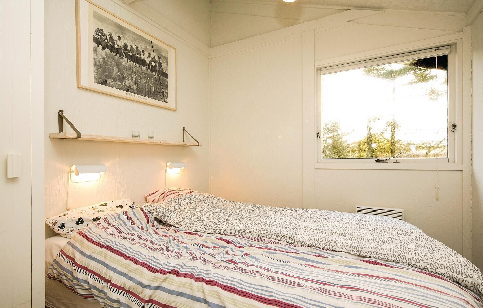 A bedroom with a bed, a window, a framed picture on the wall, and a shelf with glasses.