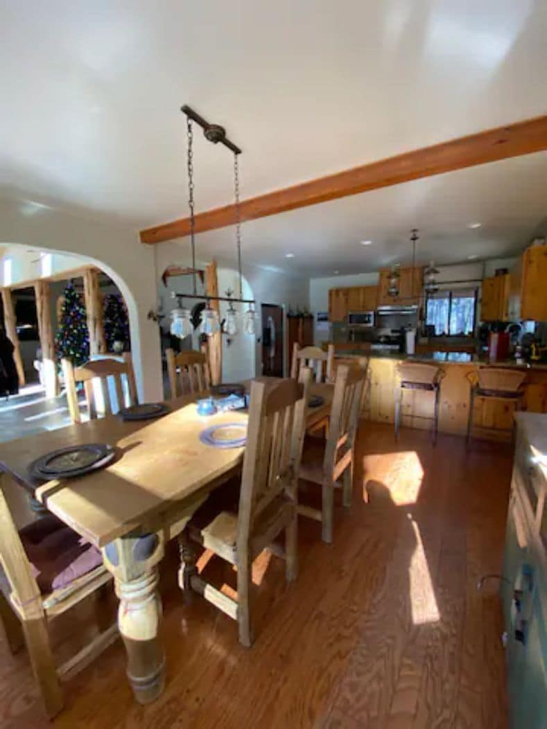 A dining area with a wooden table and chairs, a hanging light fixture, and a kitchen in the background.