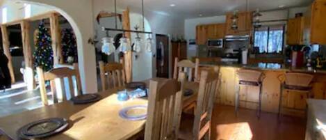 A dining area with a wooden table and chairs, a hanging light fixture, and a kitchen in the background.