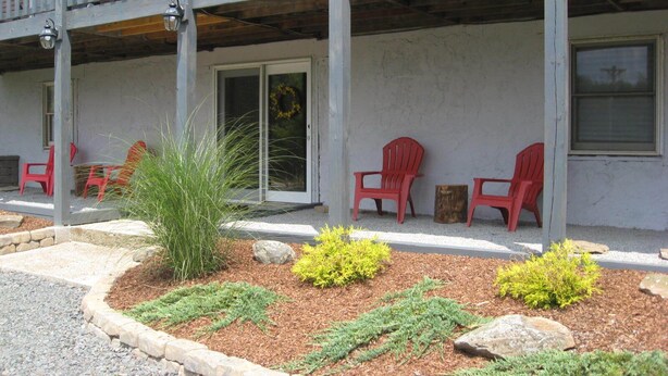 Ein Gebäude mit überdachter Veranda, roten Stühlen und einem bepflanzten Garten mit Sträuchern und Steinen.