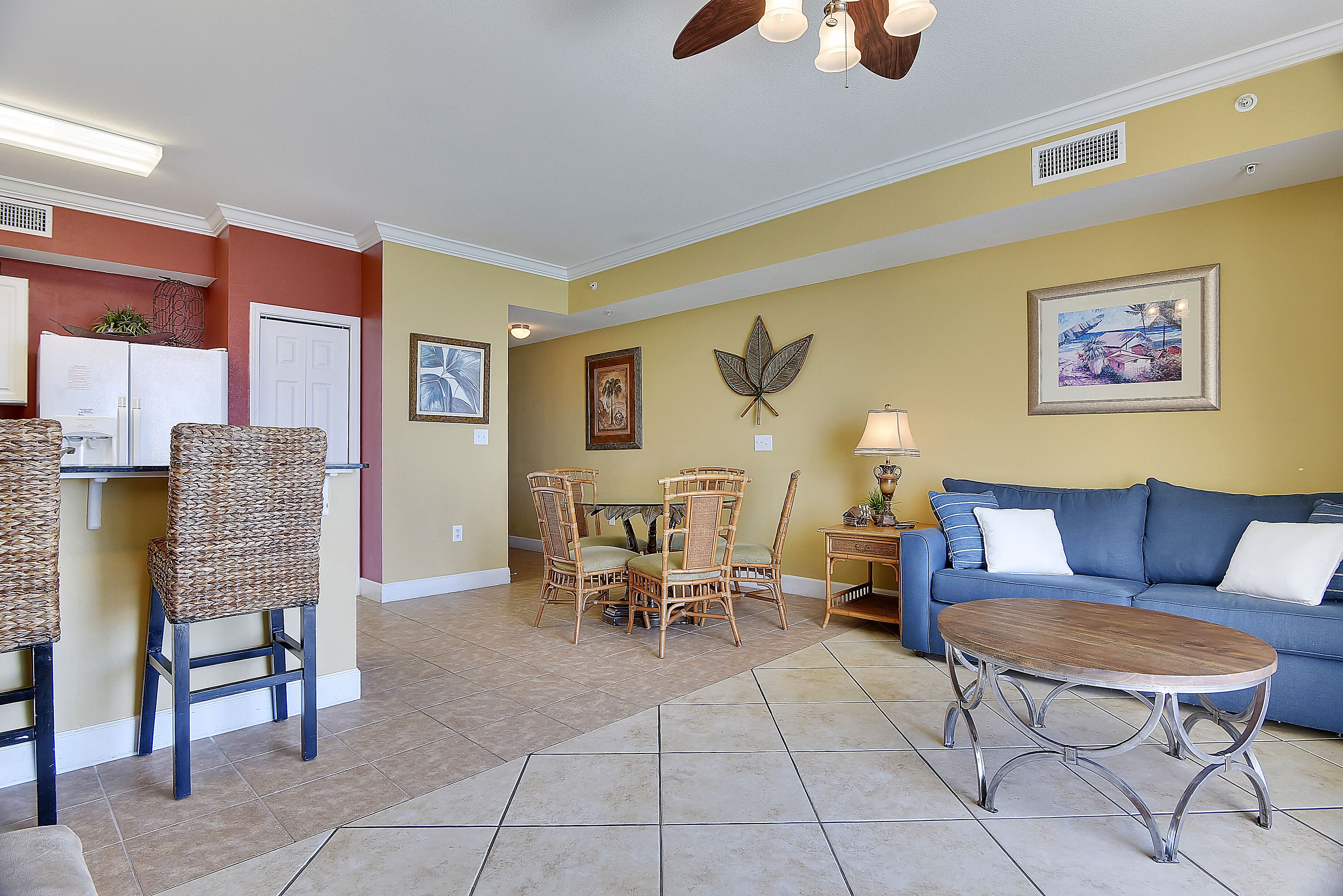 A living room with a blue sofa, wicker chairs, a wooden coffee table, and a dining area with a white refrigerator and wicker chairs.