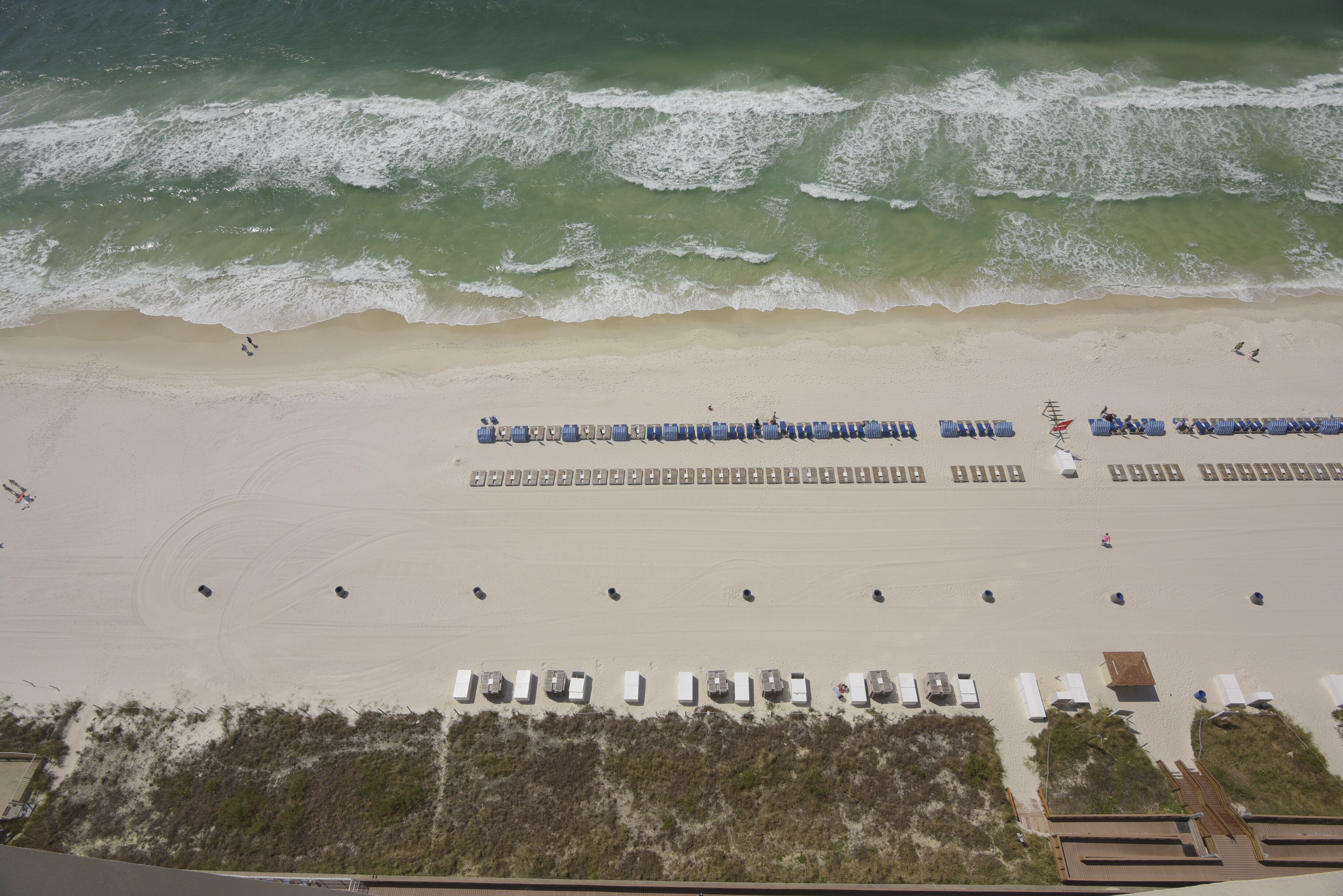 Une vue aérienne d’une plage avec des rangées de chaises longues et de parasols, une plage de sable et des vagues qui déferlent sur le rivage.