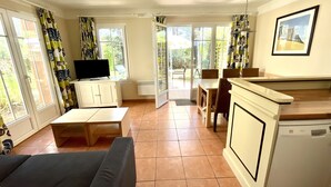 A living room with a sofa, a TV on a cabinet, a dining area with chairs and a table, and a kitchen with a dishwasher.