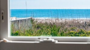 Blick durch ein Fenster auf einen Strand mit Zaun und Vegetation im Vordergrund.