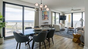 Un salon spacieux avec une grande table en verre, des fauteuils gris, un lustre moderne, un téléviseur à écran plat et un balcon offrant une vue sur l’océan.