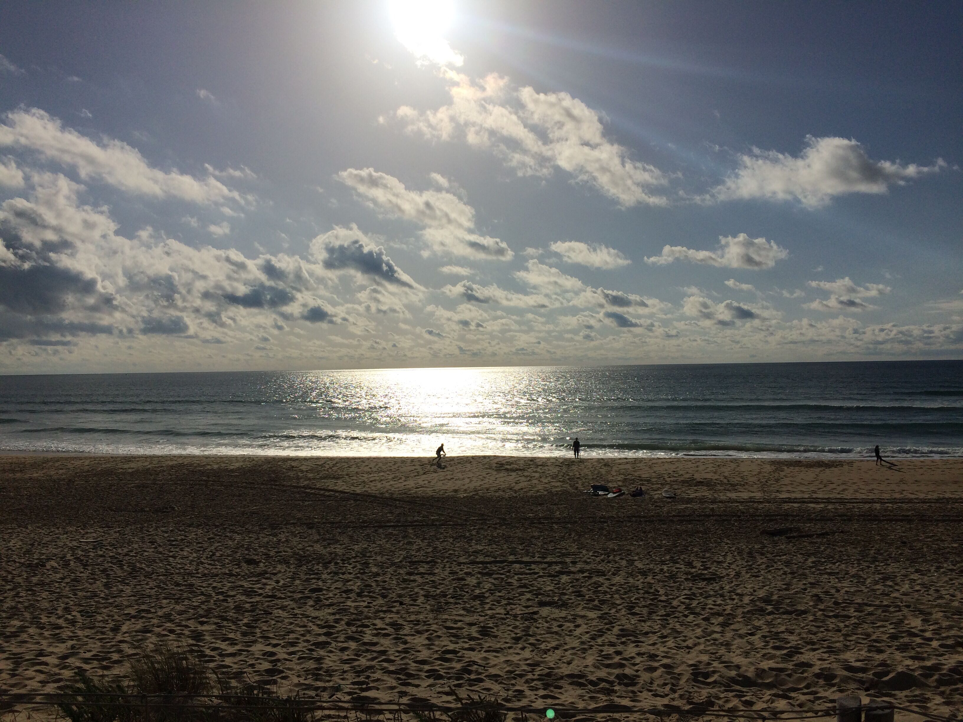 A beach with the sun reflecting off the water, a few people walking, and a clear sky.