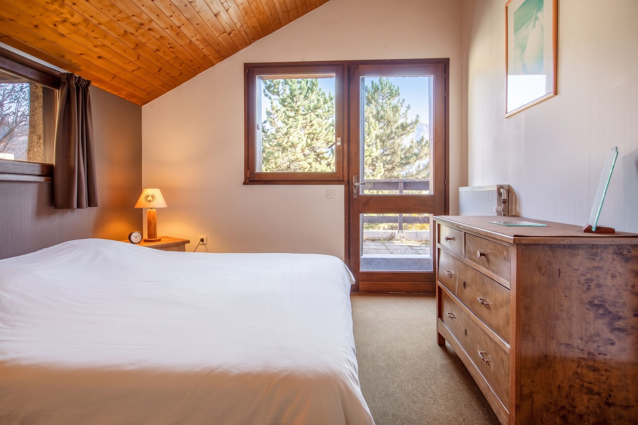 A bedroom with a bed, a dresser, a lamp, and a window with a view of trees.
