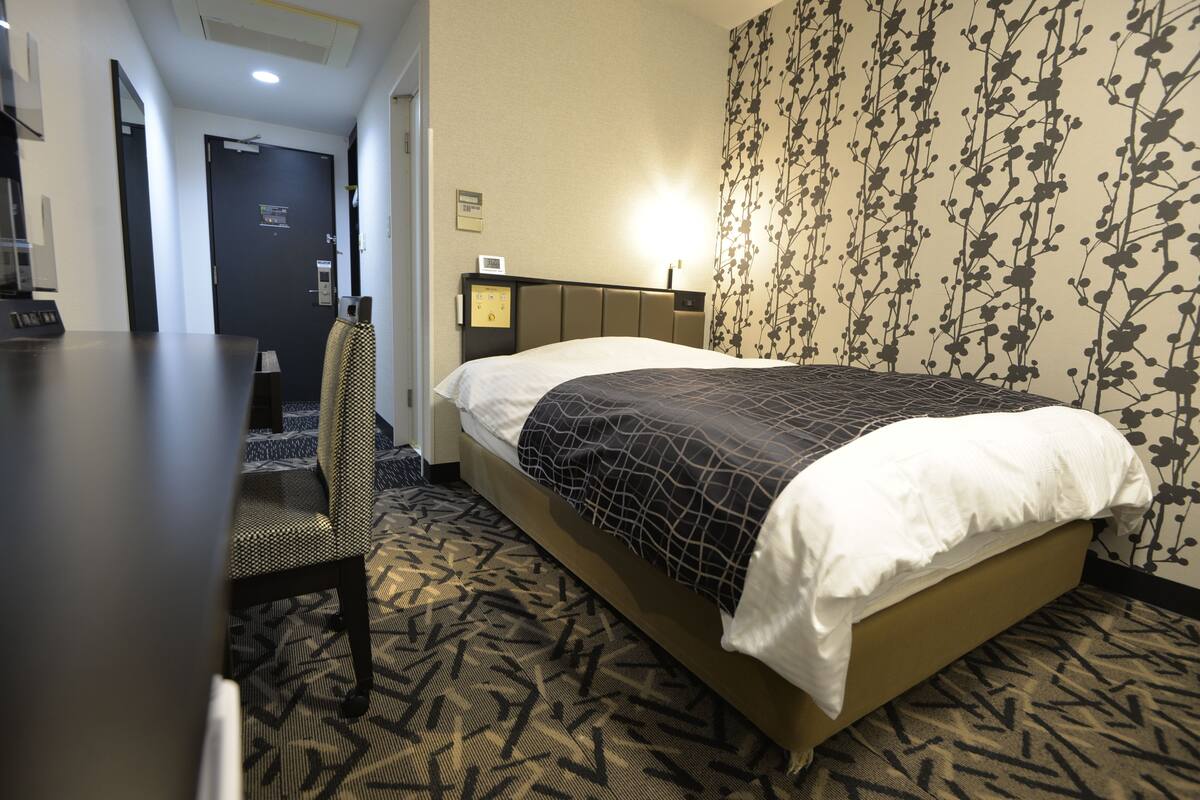 A hotel room with a bed, a desk, and a chair. There is a door and a wall with a floral pattern.